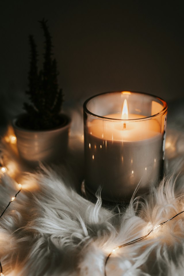 A lit candle in a glass jar beside a small plant and fairy lights